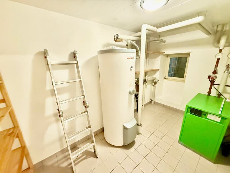 Utility room with white tiled floor, white walls, central heating system, gas meter, water tank, shelves, ladder, green electrical appliance