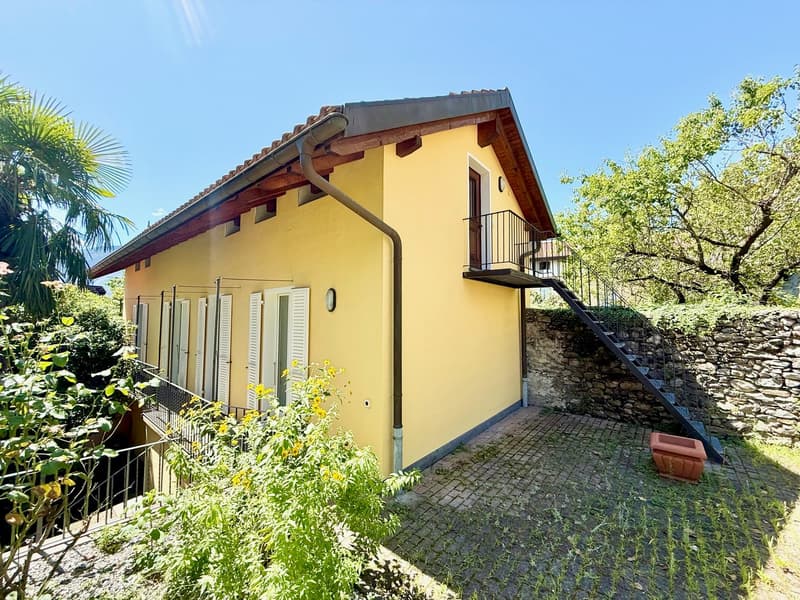 single-story yellow house with brick pavement entrance, metal staircase, balcony with railings, stone wall, large front windows with white shutters, gutter, plants and trees in the front yard