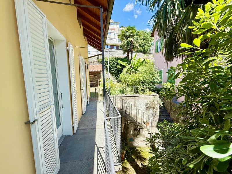 House exterior with yellow walls, white shutters, tiled pathway, metal railings, garden with plants and trees