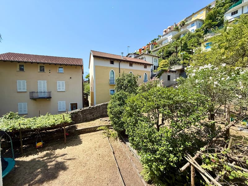 Two houses with brown roofs and white walls, balcony on the upper floor of the second house, plants and trees, visible hilly terrain