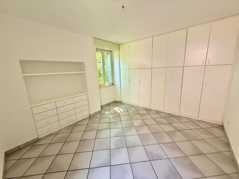 Room with white tiled floor, white walls, row of tall white wardrobes, row of white drawers, open window with greenery outside.