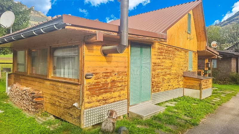 small wooden house, slanted brown roof, wooden wall, window with white curtain, stacked wood next to the house, pathway in front of the house