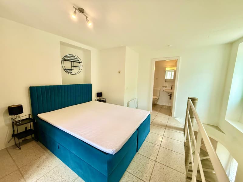 modern bedroom, teal bed, white walls, white bed linen, bedside table with lamp, open doorway leading to bathroom