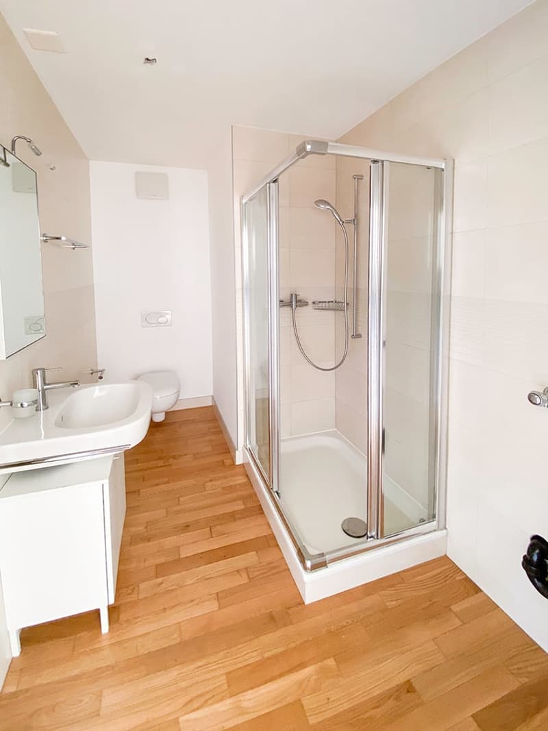 modern bathroom with wooden floor, white walls, glass shower, white sink, toilet, mirror, and cabinet