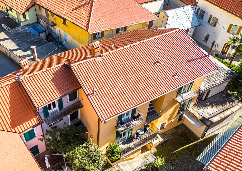 Aerial view of a yellow house with tiled roof, multiple balconies, multiple windows, and a chimney. Trees are visible around the house, and a palm tree can be seen on the side.