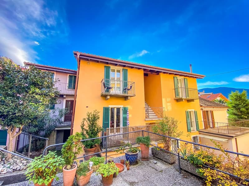 Two-story house with yellow exterior, green shutters, multiple balconies, garden with various potted plants and a tree, gravel pathway leading up to the entrance