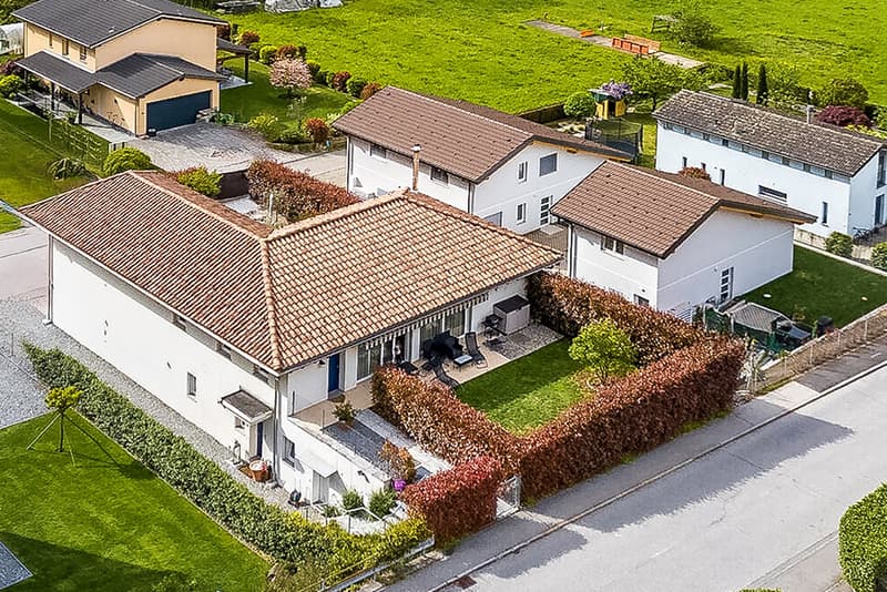 Two-story residential building with a red tile roof, surrounded by a well-maintained garden with lush greenery. The property also includes a garage and a parking area.