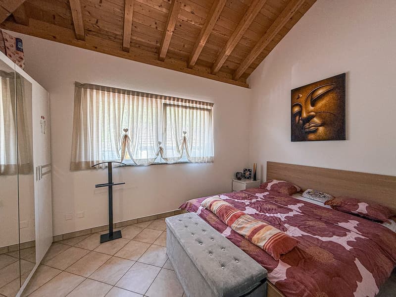Spacious bedroom with wooden ceiling, large windows, floral patterned bedding, gray tufted bench, and a decorative Buddha artwork on the wall.