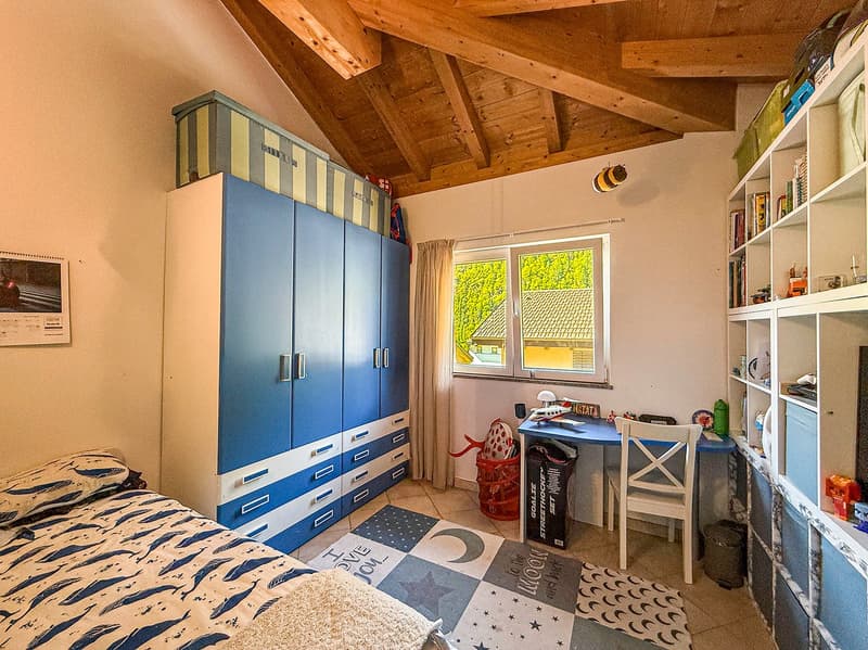 Spacious bedroom with wooden ceiling beams, large blue wardrobe, desk, shelves, and patterned rug on the floor