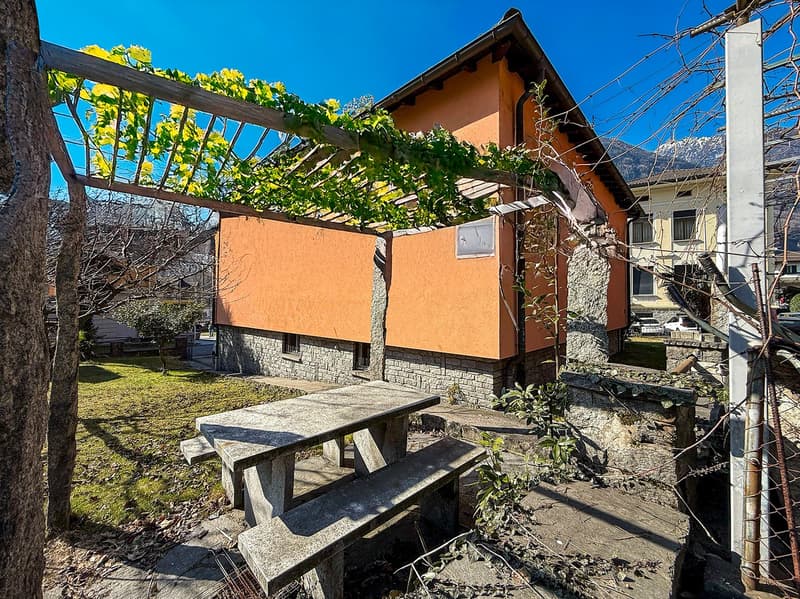A small single-story house with an orange wall and a wooden structure with vines. There is a wooden table and benches in front of the house.