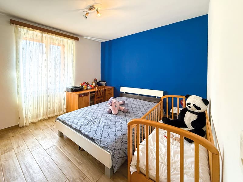 Spacious bedroom with blue accent wall, wooden furniture including a dresser, bed with patterned bedding, and stuffed animal toys including a panda bear