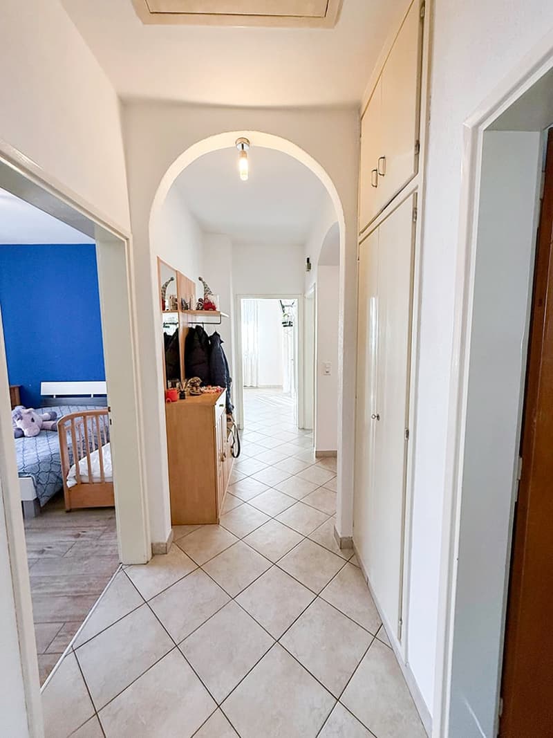 The image shows a hallway with an arched entryway, tile flooring, and a storage cabinet or closet visible in the background. The hallway leads to other rooms, including what appears to be a bedroom visible through the open doorway.