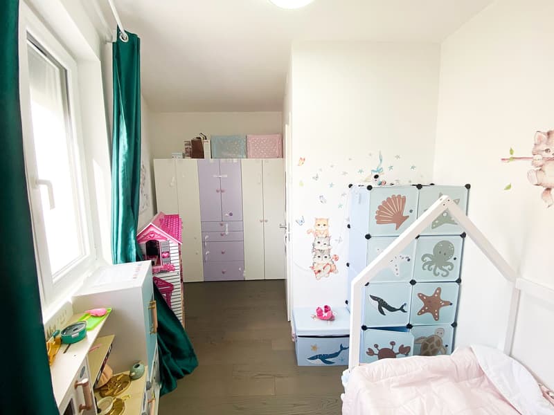 Wooden floor, white walls, various toys and storage boxes, bed with pink bedding, colorful storage unit, window with curtain
