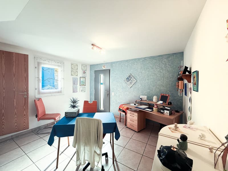 Office room with desk, office chair, plants, wooden door, tiled floor, mounted frames on the wall, white ceiling, blue desk cloth