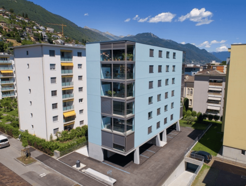 Multi-story apartment building with balconies, located in a mountainous area with scenic views. The building has a modern, minimalist design with a mix of white and gray colors.