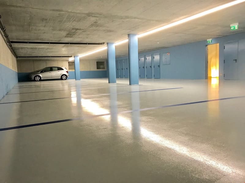 This image shows an indoor parking garage with a concrete floor, blue walls, and lockers along the side. There is a single car parked in the garage, and the space appears to be well-lit with overhead lighting.