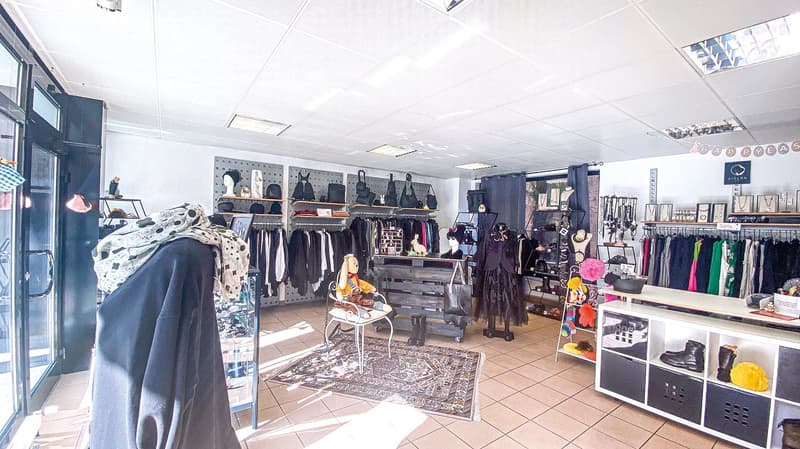 A clothing shop with rows of clothes on racks, a desk with a handbag, a small stool with a doll, a white desk with shelves, glass door, tiled floor.