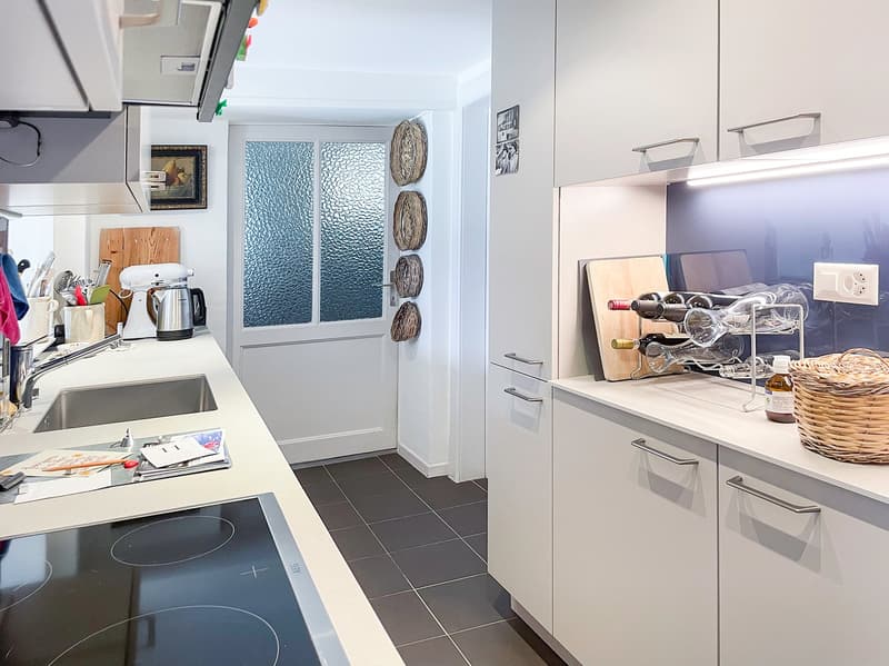 Modern kitchen, white cabinets, black tiled floor, induction hob, stainless steel sink, wine rack, white walls, glass door, countertop with basket and wine bottles