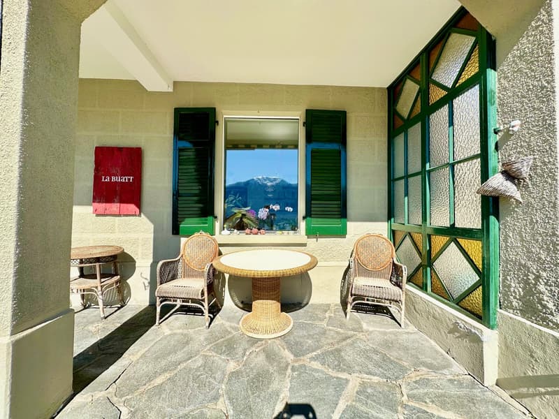 Small stone patio with wicker furniture, table and chairs, stone wall, red sign, windows with shutters, green frames