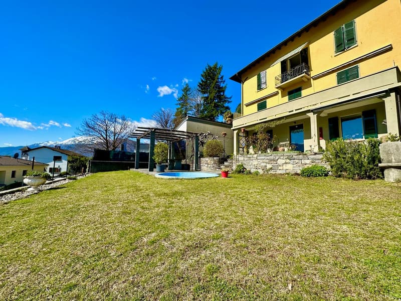 A spacious garden with a swimming pool in the middle and a stone wall with plants. The background shows a house with balconies and windows. The sky is blue with some clouds, and in the distance, there are houses and trees against the backdrop of mountains