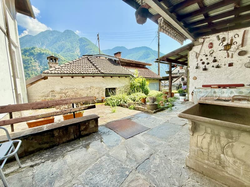 terrace with stone floor, covered by wooden awning, stone washbasin, wooden seat, various plants, metal grate on floor, mountains in the background