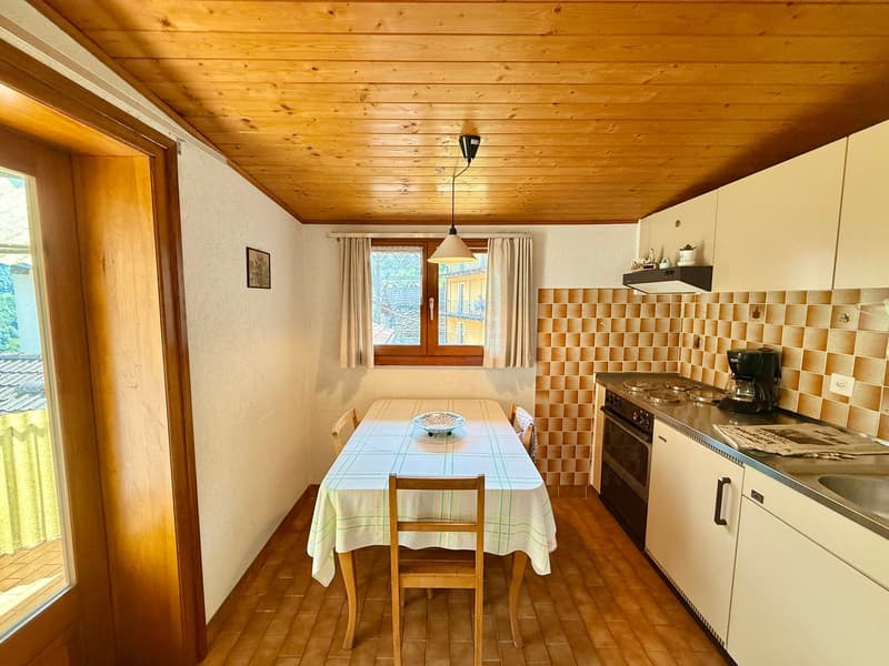 Wooden ceiling, table with chairs, sink, oven, counter with coffee maker, plate on table, window with curtains, picture frame on wall, lamp, wooden floor, kitchen cabinets
