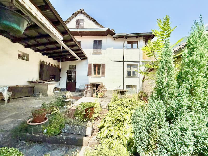 Traditional house with exposed stone walls, large wooden awning, wooden shelves, garden with various plants and trees, stone flooring