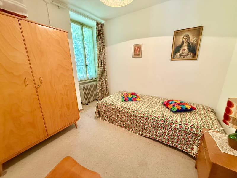 A bedroom featuring a single bed with patterned bedding, decorative pillows, and wooden wardrobe with a night light on top. The room includes a nightstand with a lamp and a small wooden chair.