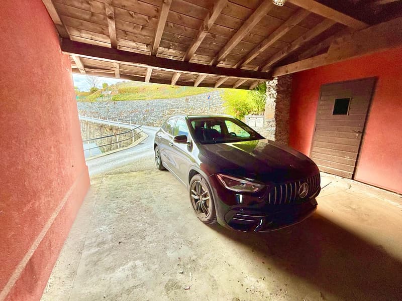 Maroon Mercedes parked in a garage beneath a wooden roof with a stone retaining wall and road