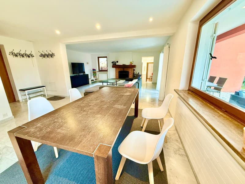 wooden table, 4 white chairs, carpet, fireplace visible in the background, flatscreen TV, couch, radiator, large window, chairs