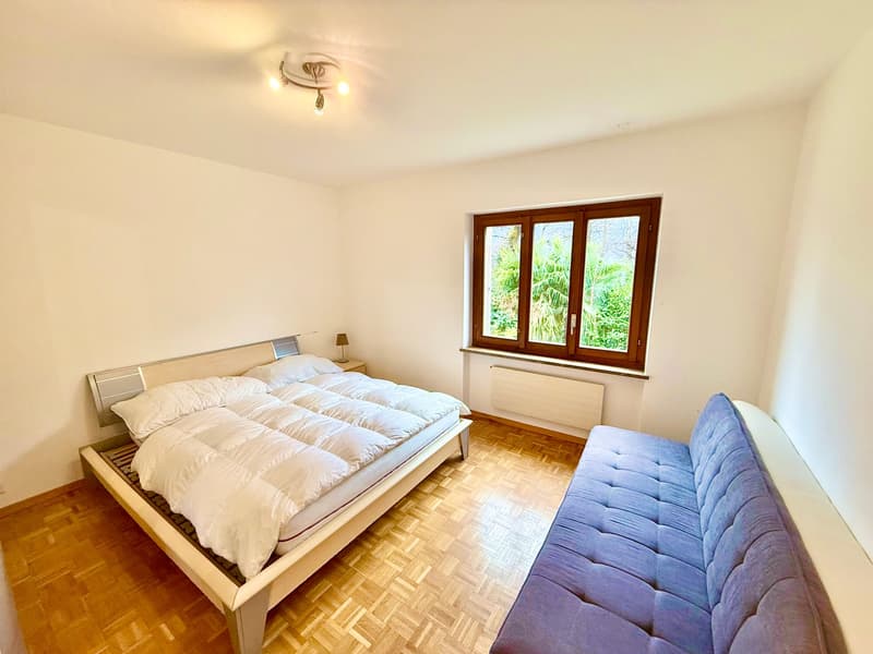 a bed with white bedding, a side table with a lamp, a window, a couch, and wooden flooring