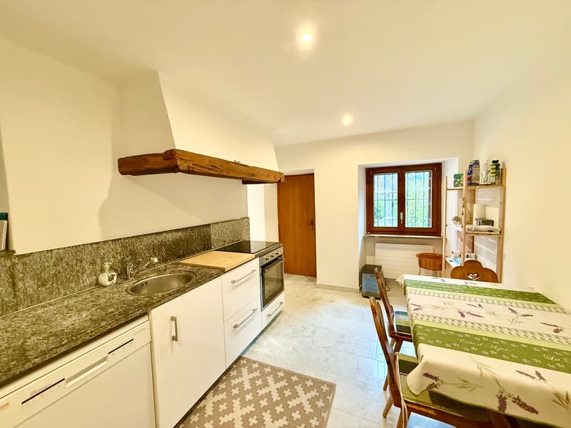 Modern kitchen with granite countertops, stainless steel sink, wooden shelves, built-in oven, dishwasher, dining table with green tablecloth, wooden chairs, wooden door, glass window, white walls, tiled floor.