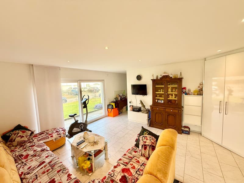 Open living room, large windows, white flooring, couch, television, table, exercise bike, wardrobe, small white cabinets