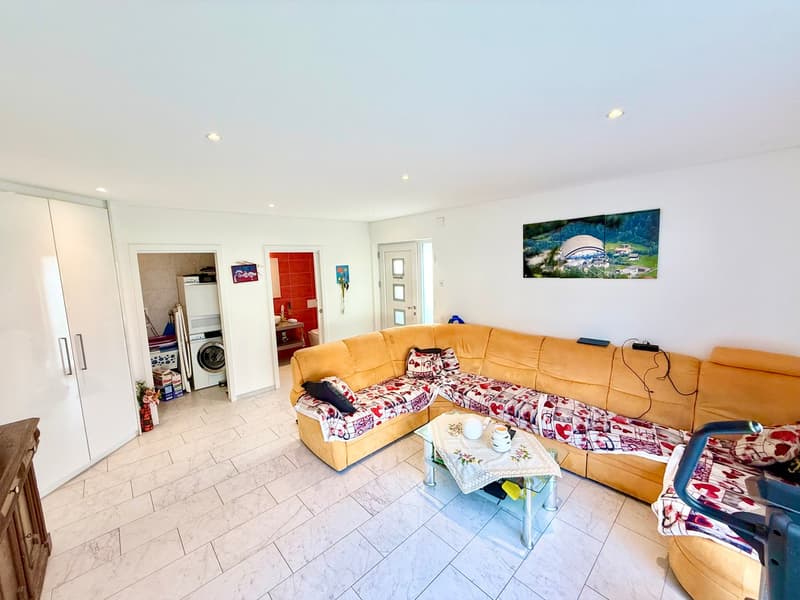 a spacious and bright living room with white tile flooring and white walls, a large beige sectional sofa, a glass coffee table, a white laundry machine in a laundry room, and a decorative painting on the wall