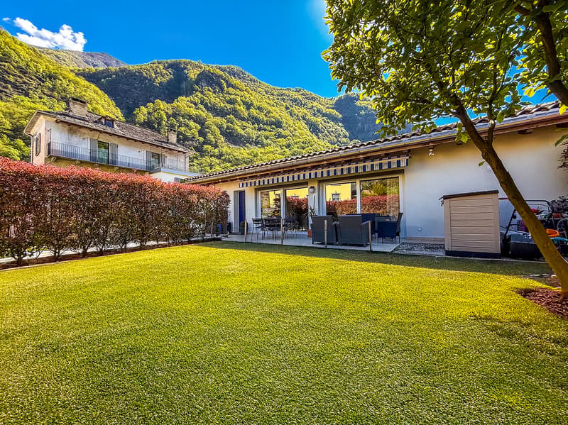 Two-story house with a balcony, surrounded by lush greenery and mountains in the background. The house has a patio area with outdoor furniture and a garage on the side.