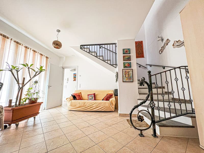 Spacious living room with tile flooring, large windows, a beige sofa, decorative wall art, and a wrought iron staircase leading to the upper level.