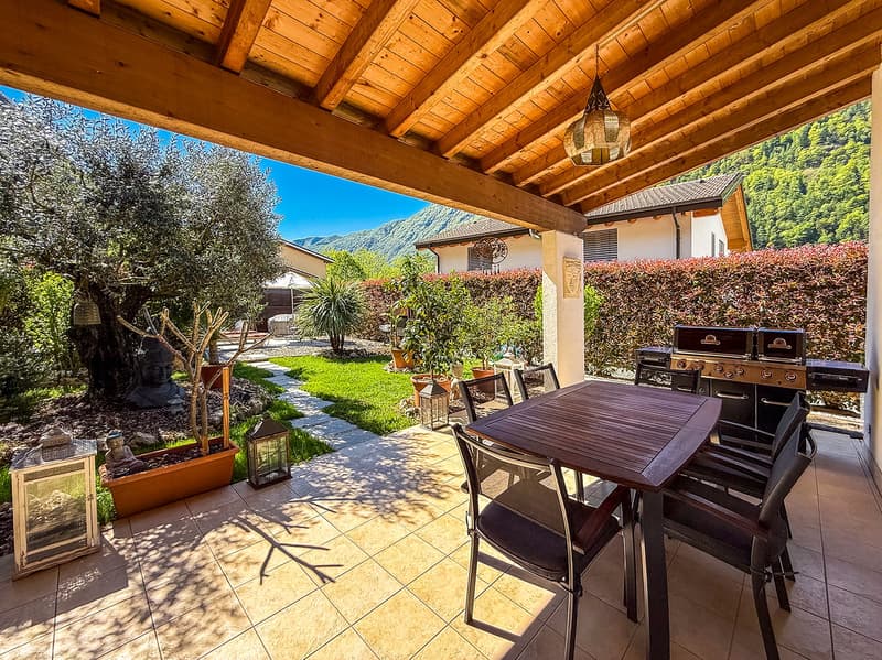 Covered patio with wooden beamed ceiling, tiled floor, outdoor dining table and chairs, barbecue grill, lanterns, and views of a lush garden and mountains in the background