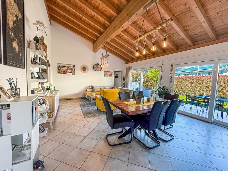 Spacious open-plan living area with wooden beamed ceiling, large windows overlooking a balcony or terrace, dining table with chairs, and shelving unit displaying decor and artwork.