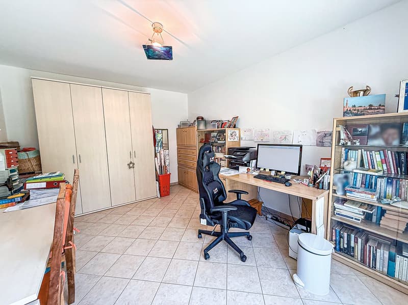 The image shows a home office space with a desk, computer, and office chair. There are bookshelves, cabinets, and various personal items and decor visible in the room. The floor is tiled and the room appears to be well-lit.