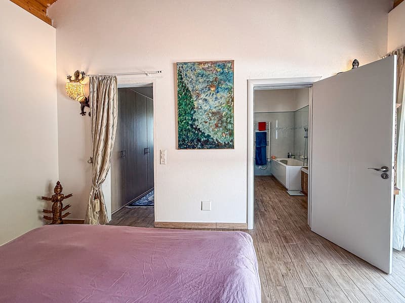 Spacious bedroom with hardwood floors, white walls, large artwork, and an open doorway leading to a bathroom