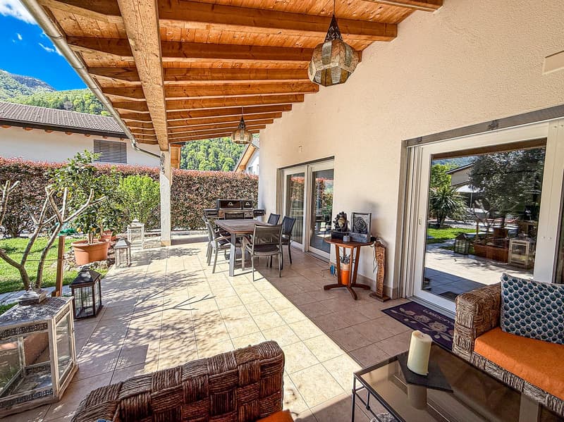 Covered patio with wooden beamed ceiling, tiled flooring, outdoor dining table and chairs, lanterns, potted plants, and views of the surrounding greenery and mountains.
