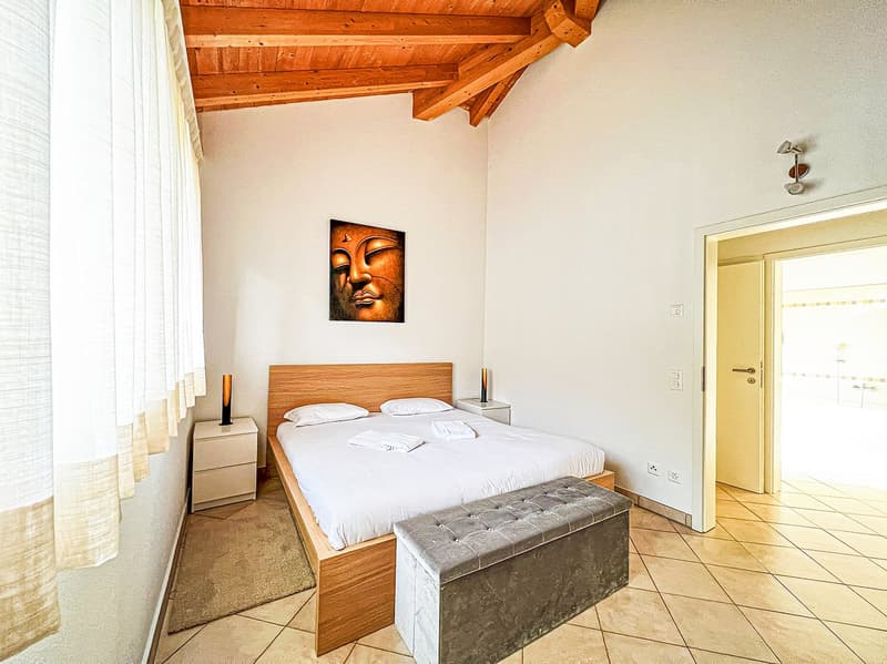 Spacious bedroom with wooden ceiling beams, a large bed with white bedding, a wooden headboard, and a Buddha artwork on the wall. The room also features a tiled floor and a stone bench at the foot of the bed.
