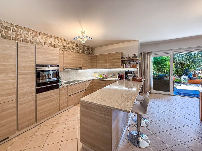 Spacious kitchen with modern wood-paneled cabinets, granite countertops, stainless steel appliances, and a sliding glass door leading to a balcony or terrace area.