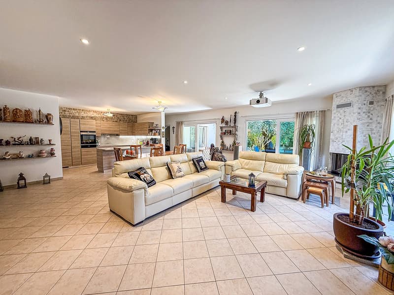 Modern living room with beige couches, beige tiled floor, open to a kitchen with a dining area, large windows, and potted plants