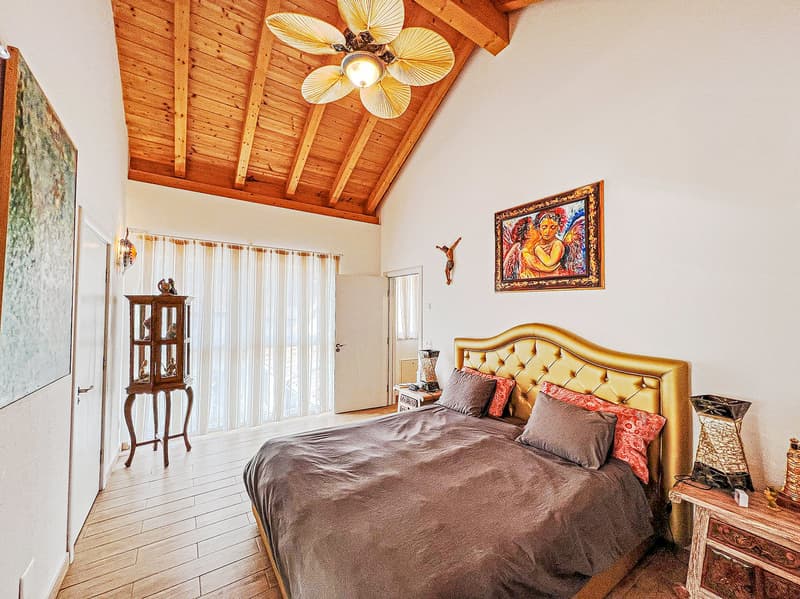 Spacious bedroom with wooden ceiling, large windows, gold-tufted headboard, wooden furniture, and a decorative painting on the wall.