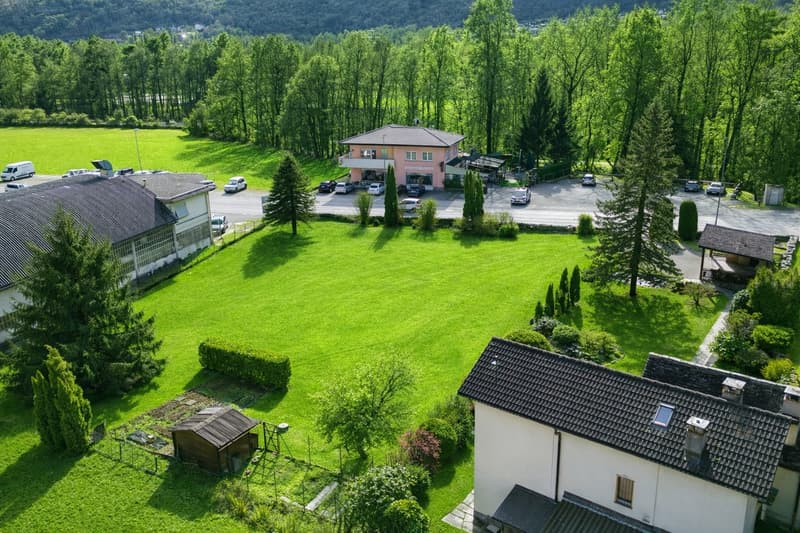 Several buildings, parking lot with multiple cars, lush green lawn, various trees, and mountainous backdrop.