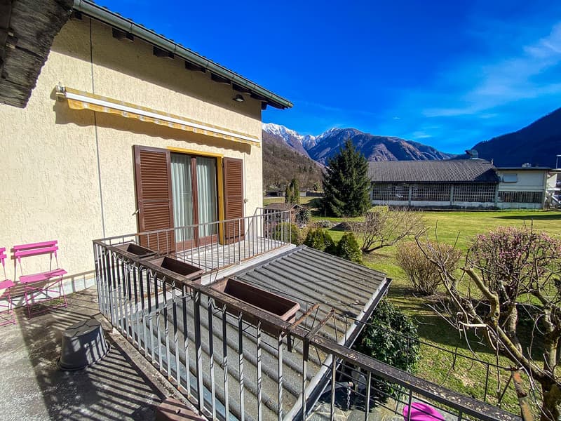 House with balcony, metal railing, awning, metal roof, pink chairs, metal planter, green grass, trees, mountains in the background