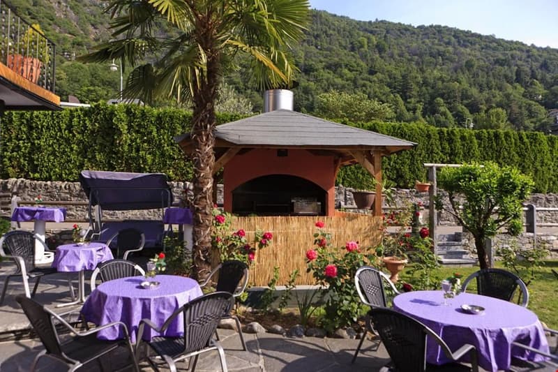Outdoor dining area with a covered gazebo, palm trees, and tables set with purple tablecloths. There is a stone fireplace in the gazebo and the area is surrounded by lush greenery and mountains in the background.