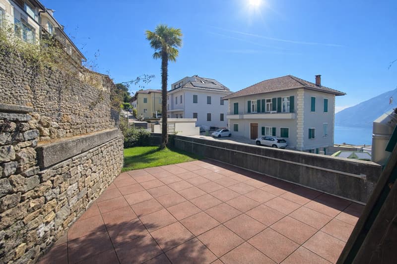 Tiled roof, white facade, multiple windows, garden, 2 cars parked in front, stone wall, clear sky, sea view