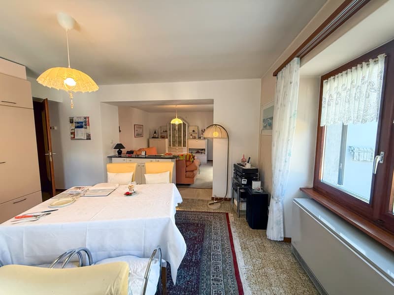 The image shows an open-plan living room and kitchen area. The living room has a white tablecloth-covered dining table, yellow chairs, and a patterned rug on the floor. The kitchen area is visible in the background, with cabinets, appliances, and a counte